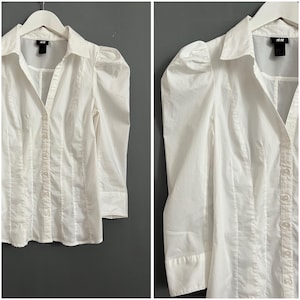 May include: White long-sleeve button-up shirt with puffed sleeves and a collar. The shirt has a single button closure at the top and a row of buttons down the front.