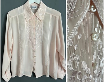 Puede incluir: Una blusa de manga larga de color rosa pálido con cierre de botones en la parte delantera y un delicado bordado floral. La blusa tiene cuello y una etiqueta que dice "Greshe".