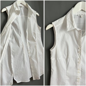 May include: White sleeveless button-down shirt with thin vertical stripes. The shirt is unbuttoned and the collar is folded down.