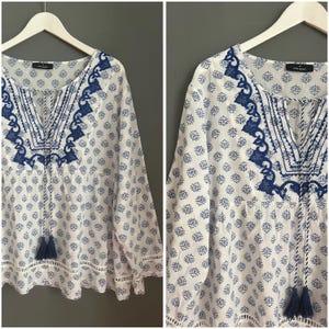 Può includere: Camicetta bianca con motivo floreale blu e finiture ricamate blu intorno allo scollo. La camicetta ha maniche lunghe e un laccio con nappe allo scollo a V. L'orlo inferiore e i polsini hanno un delicato bordo in pizzo.