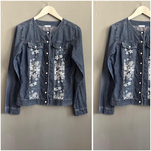 Puede incluir: Chaqueta vaquera azul con cuello redondo y mangas largas. La chaqueta presenta diseños florales blancos en la parte delantera y se abrocha con botones. Dos pequeños bolsillos en el pecho.