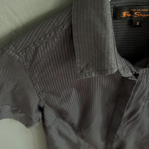 May include: A grey short-sleeved shirt with a vertical striped pattern. The shirt has a collar and a button closure. The brand label "Ben Sherman" is visible on the collar. The shirt is size small.