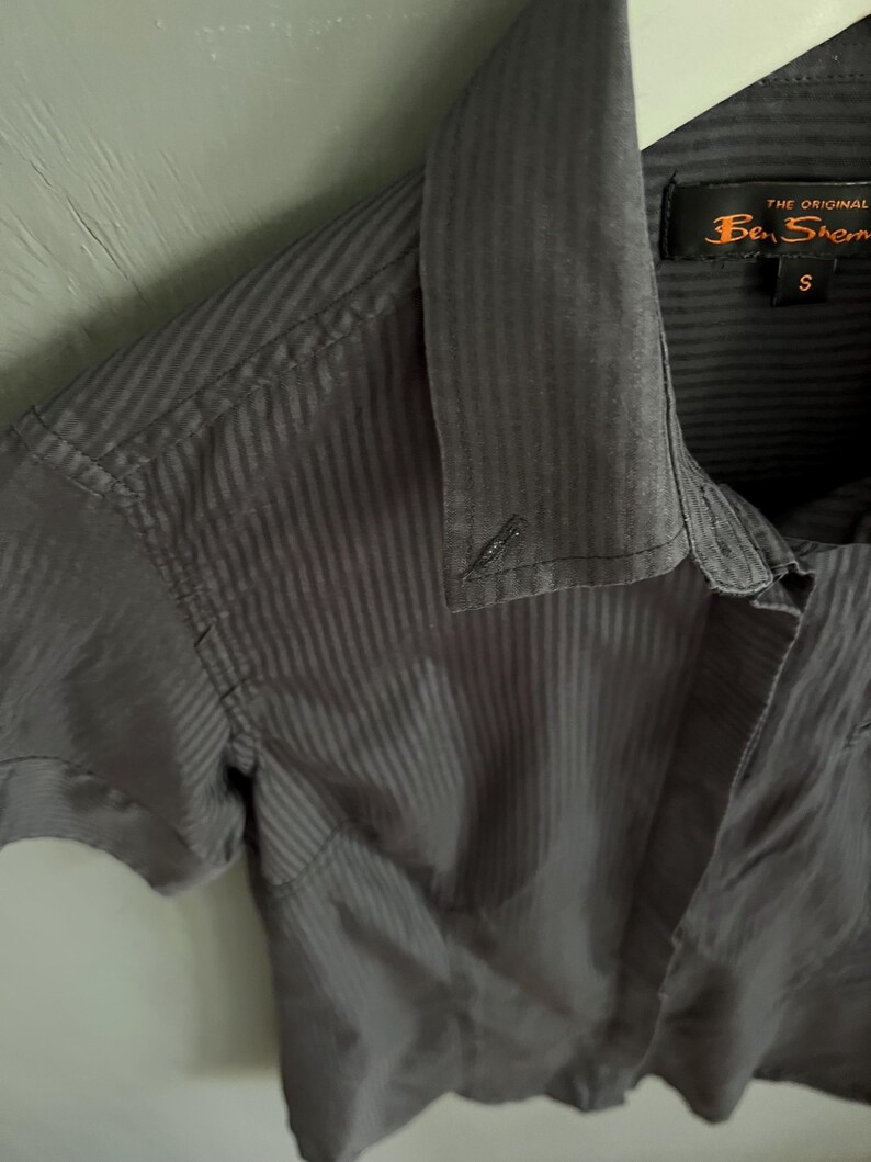 May include: A grey short-sleeved shirt with a vertical striped pattern. The shirt has a collar and a button closure. The brand label "Ben Sherman" is visible on the collar. The shirt is size small.