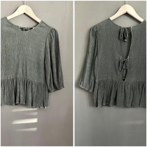 Pode incluir: Uma blusa verde sálvia com tecido texturizado e plissado. A parte superior apresenta mangas três quartos, decote redondo e bainha com babados. A parte de trás tem uma abertura em forma de fechadura com fechos de laço. A blusa está pendurada em um cabide de madeira.