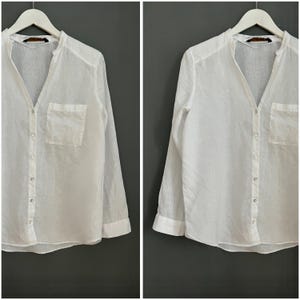May include: White linen button-down shirt with a v-neck and a single pocket on the left side.