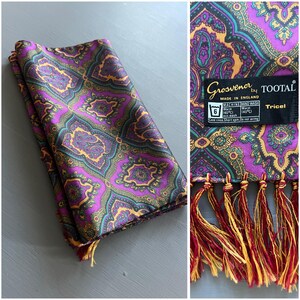 May include: A folded purple paisley scarf with gold, teal, and black accents. The scarf has a decorative fringe and a tag that reads "Grosvenor by TOOTAL, Made in England". The scarf is made of Tricel.