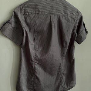 May include: A grey short-sleeved shirt with vertical stripes. The shirt features a collar and cuffed sleeves. It is hanging on a white hanger against a grey wall.