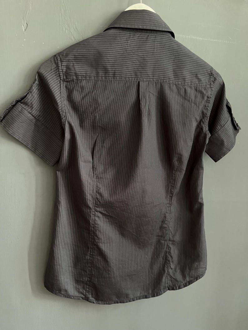 May include: A grey short-sleeved shirt with vertical stripes. The shirt features a collar and cuffed sleeves. It is hanging on a white hanger against a grey wall.