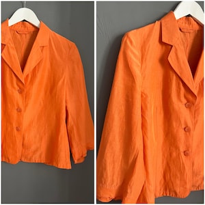 Puede incluir: Una chaqueta de color naranja brillante con cuello clásico y tres botones en la parte delantera. La chaqueta tiene mangas largas con una ligera forma de campana en los puños.