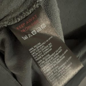 May include: A dark gray fabric tag with care instructions and the text "KEEP AWAY FROM". The tag includes washing, ironing, and drying symbols, along with the fabric composition and country of origin.