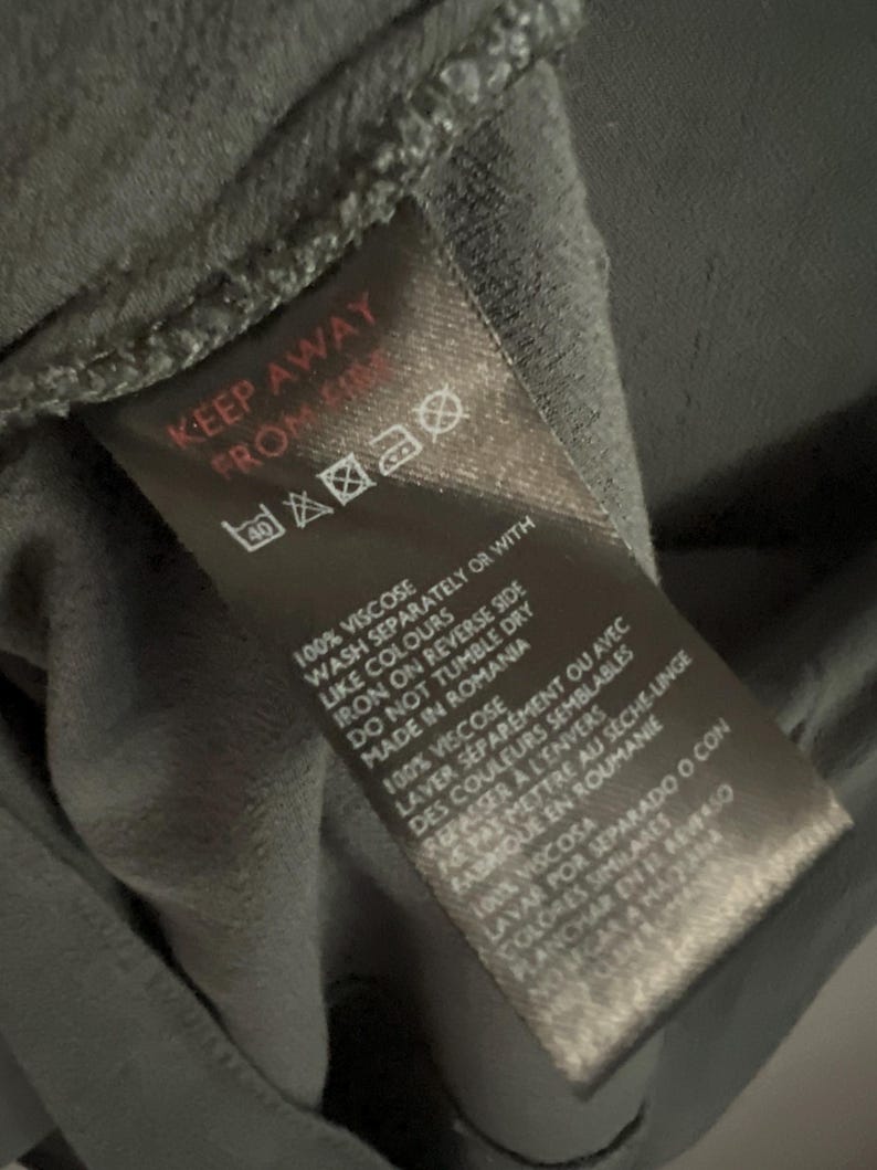 May include: A dark gray fabric tag with care instructions and the text "KEEP AWAY FROM". The tag includes washing, ironing, and drying symbols, along with the fabric composition and country of origin.