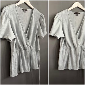 May include: A light blue wrap top with short, puffed sleeves and a peplum hem. The top has a V-neckline and a tie closure at the waist. The top is hanging on a white hanger against a gray background.