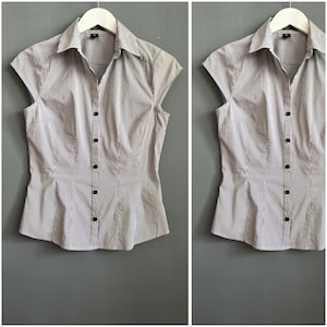 May include: A grey and white striped button-down shirt with short sleeves. The shirt has a collared neckline and black buttons. The shirt is hanging on a white hanger against a grey background.