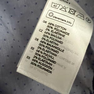 Op de afbeelding: Close-up van een kledinglabel met tekst in meerdere talen. Het label vermeldt de stof samenstelling als katoen, polyamide en elastaan. Het label bevat ook wasvoorschriften en de tekst "clevercare.info".
