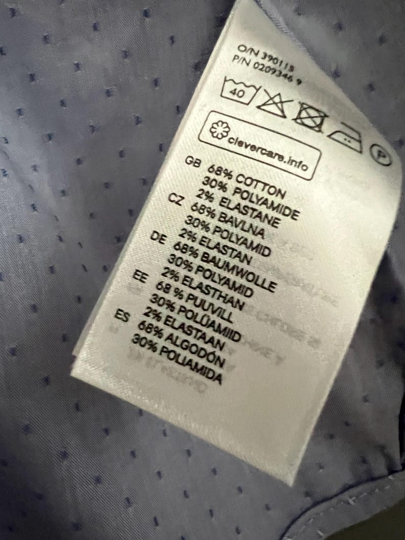 Op de afbeelding: Close-up van een kledinglabel met tekst in meerdere talen. Het label vermeldt de stof samenstelling als katoen, polyamide en elastaan. Het label bevat ook wasvoorschriften en de tekst "clevercare.info".