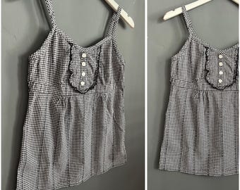 May include: A black and white gingham print camisole top with thin straps. The top features a ruffled detail around the neckline and a row of white buttons down the center. The garment is hanging on a white hanger against a gray background.