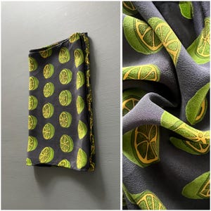 Puede incluir: Tela gris oscuro doblada con un patrón repetido de rodajas de lima verdes y amarillas. La tela tiene una textura suave y un ribete dorado. La imagen también muestra un primer plano de la tela, destacando los detalles del diseño de lima.
