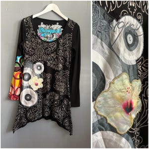 Op de afbeelding: Zwarte tuniek met lange mouwen en een uniek design. De tuniek heeft een bloemen- en abstract patroon, een kleurrijke mouw en een bloemenapplicatie. Het woord "Desigual" staat op de halslijn.