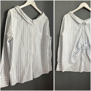 Può includere: Una camicia a maniche lunghe a righe bianche e blu con chiusura a bottoni sul davanti e un dettaglio a fiocco sul retro.