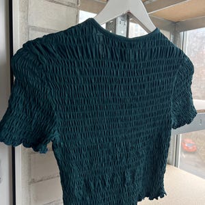 K&ouml;nnte beinhalten: Ein dunkel t&uuml;rkisfarbenes Oberteil mit kurzen &Auml;rmeln und einem strukturierten, ger&uuml;schten Design. Das Oberteil h&auml;ngt an einem wei&szlig;en Kleiderb&uuml;gel. Der Stoff hat ein geripptes Aussehen, und die &Auml;rmel haben einen gewellten Rand. Das Oberteil ist aus einem dehnbaren Material.