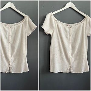 Pode incluir: Blusa creme de manga curta, com decote redondo e botões frontais. O tecido tem uma aparência sutilmente texturizada e está pendurada em um cabide branco sobre um fundo cinza.