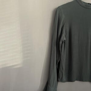 May include: A long-sleeved, olive green blouse with a high neckline and flared cuffs. The blouse is hanging on a white hanger against a gray wall. The fabric appears to be lightweight and flowing.