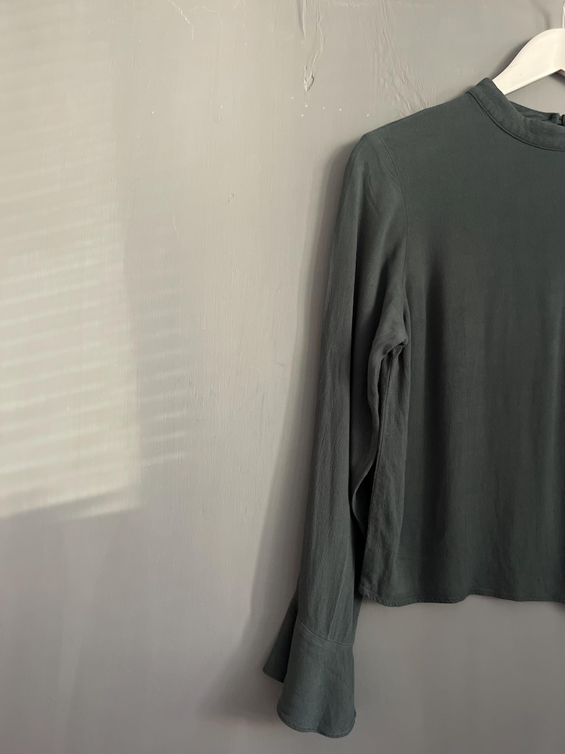 May include: A long-sleeved, olive green blouse with a high neckline and flared cuffs. The blouse is hanging on a white hanger against a gray wall. The fabric appears to be lightweight and flowing.