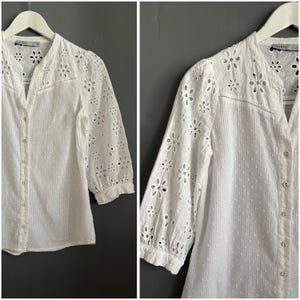 May include: White button-down blouse with floral cut-out details on the sleeves and yoke. The blouse has a mandarin collar and three-quarter sleeves. The fabric has a subtle dotted texture. The brand name "Oasis" is visible on the label.