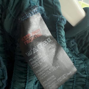 K&ouml;nnte beinhalten: Nahaufnahme eines dunkelblauen Kleidungsst&uuml;cks mit ger&uuml;schtem Design. Ein schwarzes Etikett zeigt Pflegehinweise und Stoffzusammensetzung: 97 % Viskose, 3 % Elasthan. Das Etikett enth&auml;lt auch den Text "KEEP AWAY FROM FIRE".