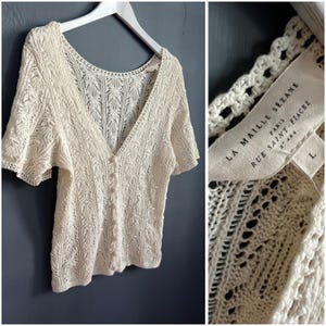 May include: An off-white short-sleeve cardigan with a V-neckline and a button-front closure. The knit fabric features an intricate openwork pattern. A label inside reads LA MAILLE SEZANE PARIS RUE SAINT-FIACRE 2E ARR. and indicates a size L.