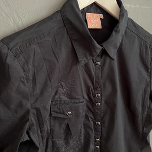 May include: Black button-down shirt with short sleeves, a chest pocket, and a collar. The shirt has a subtle, faded design on the front and a tag with the word "PACTH" on the collar.