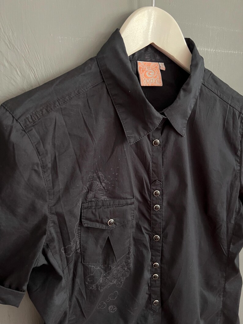 May include: Black button-down shirt with short sleeves, a chest pocket, and a collar. The shirt has a subtle, faded design on the front and a tag with the word "PACTH" on the collar.