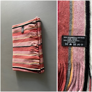 May include: Folded scarf with horizontal stripes in shades of pink, coral, and cream, with black and gray accents. The scarf has fringed edges and a label with text. The scarf is made of 50% new wool and 50% acrylic.