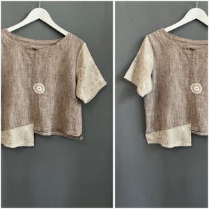 May include: A brown linen top with short sleeves and a round neckline. The sleeves are a light beige color with a floral pattern. A small, white, crocheted flower is centered on the chest. The bottom hem is a light beige color with a floral pattern.