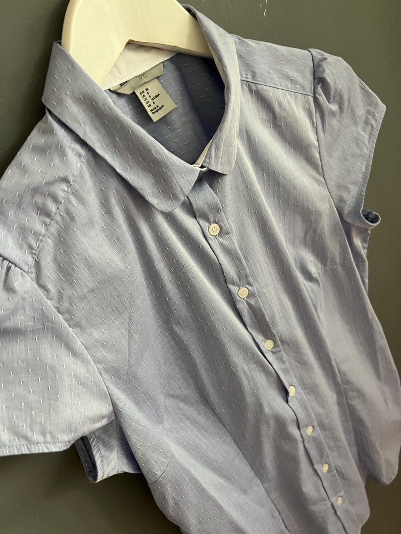 Op de afbeelding: Een lichtblauw overhemd met korte mouwen en een witte kraag. Het overhemd is voorzien van knopen en heeft een subtiel stippenpatroon. De knopen zijn wit.