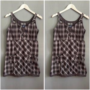 May include: Brown and white plaid sundress with adjustable straps and button details. The dress features two front pockets with bow accents and is displayed on a wooden hanger. The dress has a scoop neckline and a brown hemline.