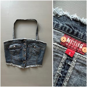 May include: A dark blue denim halter top featuring a button-down front, two chest pockets, and frayed edges along the neckline and hem. The interior label displays the text Mustang Jeans with a size S tag attached to the red fabric label.
