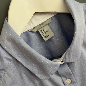 Op de afbeelding: Een lichtblauw overhemd met een witte kraag en kleine witte stippen. Het overhemd hangt aan een witte hanger. Het label luidt "H&M".