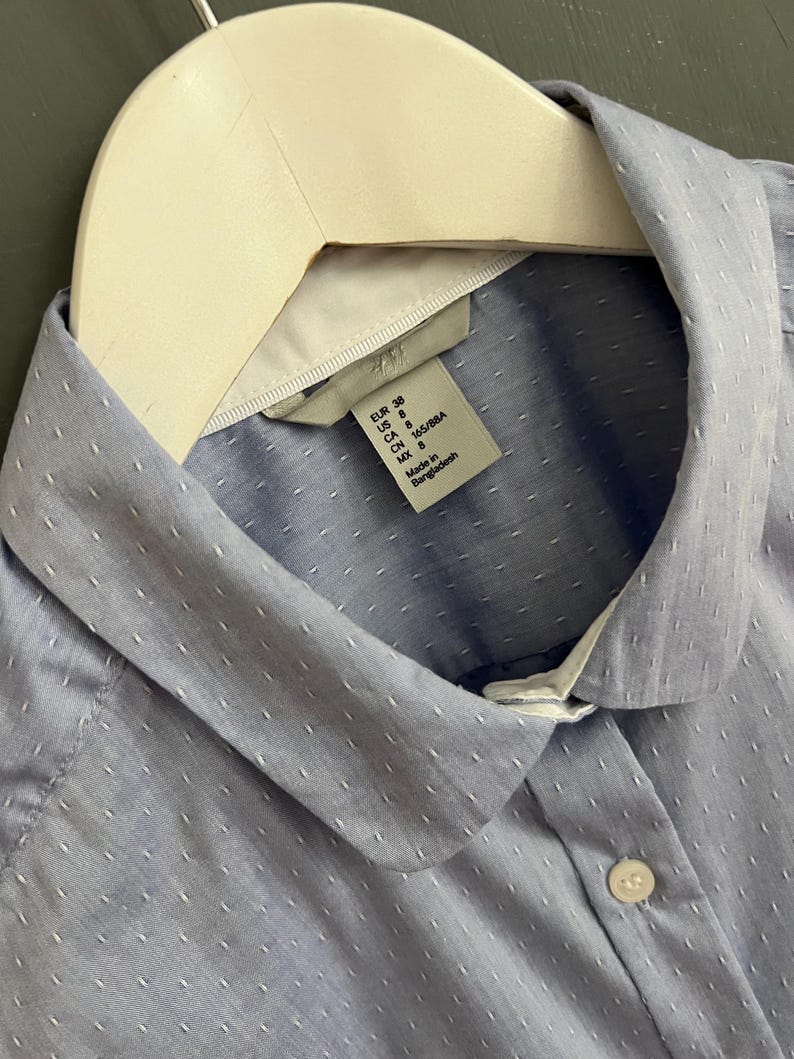 Op de afbeelding: Een lichtblauw overhemd met een witte kraag en kleine witte stippen. Het overhemd hangt aan een witte hanger. Het label luidt "H&M".