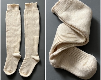 May include: Two pairs of off-white, knee-high socks. The socks are made of a thick, ribbed knit material. One pair is laid out flat, while the other is folded. The socks have a ribbed cuff at the top and a reinforced heel and toe.