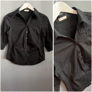 Puede incluir: Camisa negra abotonada con mangas tres cuartos. La camisa presenta bordados florales en la parte superior izquierda del pecho y un detalle de cintura fruncida. La camisa está colgada de una percha blanca.