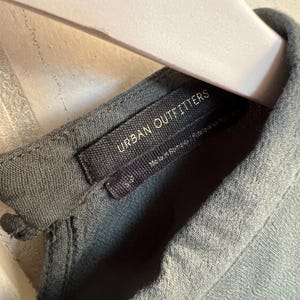 May include: Close-up of a gray-green garment tag with the text "URBAN OUTFITTERS" in white. The tag is attached to a matching fabric. The garment is hanging on a light-colored hanger.