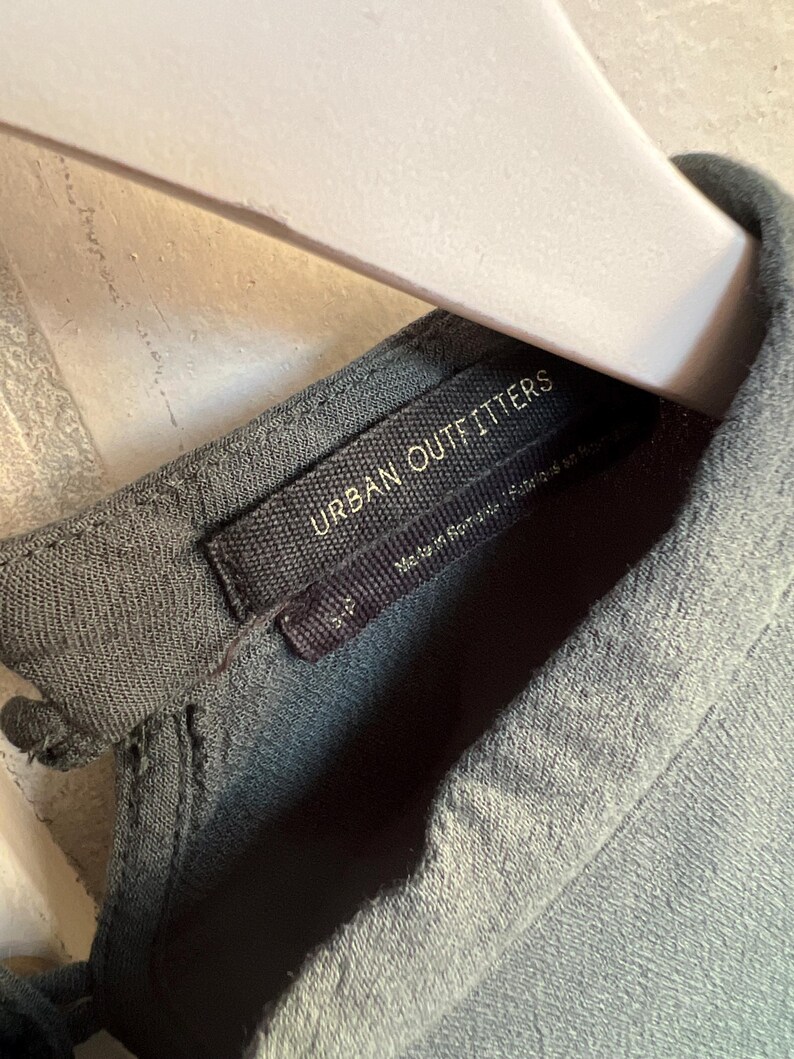 May include: Close-up of a gray-green garment tag with the text "URBAN OUTFITTERS" in white. The tag is attached to a matching fabric. The garment is hanging on a light-colored hanger.
