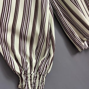 May include: Close-up of a green and brown striped long-sleeve shirt with a gathered elastic cuff. The shirt has a subtle dotted pattern.