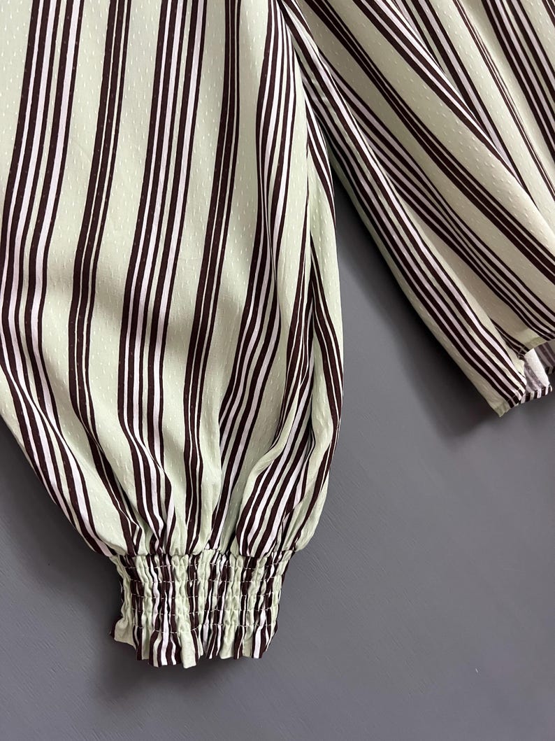 May include: Close-up of a green and brown striped long-sleeve shirt with a gathered elastic cuff. The shirt has a subtle dotted pattern.