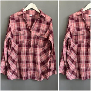 Può includere: Una camicia a quadri a maniche lunghe nei toni del bordeaux, rosa e crema. La camicia presenta un colletto, due tasche sul petto e una paramontura con bottoni. Il tessuto ha un aspetto morbido e leggermente usurato.