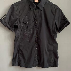 May include: Black short-sleeve button-down shirt with a collared neckline. The shirt features a small pocket on the left chest and roll-up sleeves. A subtle floral design is visible on the left side of the shirt.