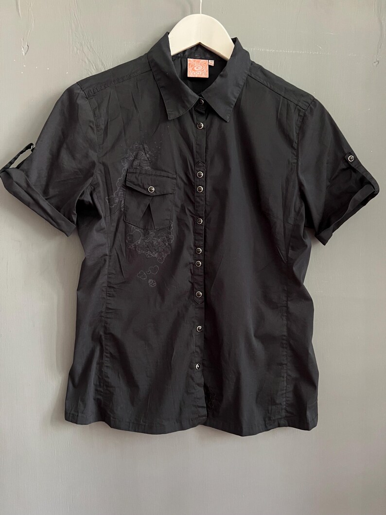 May include: Black short-sleeve button-down shirt with a collared neckline. The shirt features a small pocket on the left chest and roll-up sleeves. A subtle floral design is visible on the left side of the shirt.