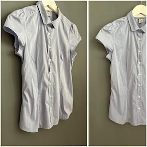 Op de afbeelding: Lichtblauw overhemd met korte mouwen en streepjespatroon. Het overhemd heeft een ronde kraag en kleine pofmouwen. Het wordt met knopen gesloten en hangt aan een witte hanger tegen een grijze achtergrond.