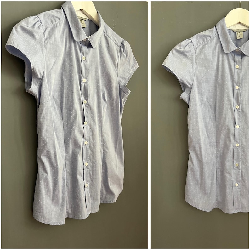 Op de afbeelding: Lichtblauw overhemd met korte mouwen en streepjespatroon. Het overhemd heeft een ronde kraag en kleine pofmouwen. Het wordt met knopen gesloten en hangt aan een witte hanger tegen een grijze achtergrond.